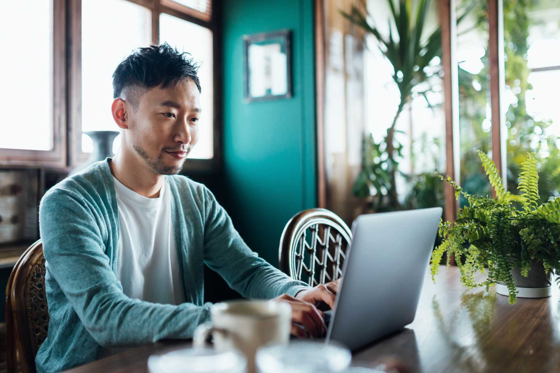 A young Asian man wearing a blue hoodie with a white t-shirt sits at his laptop.
