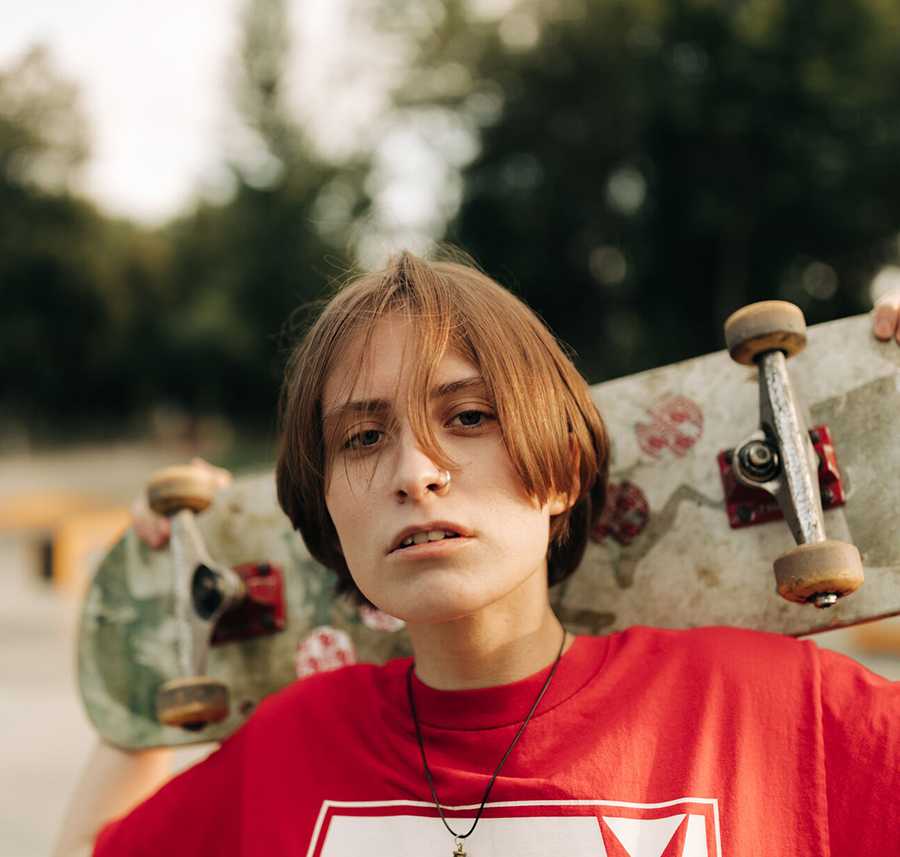 A genderfluid young person carries a skateboard on their shoulders.