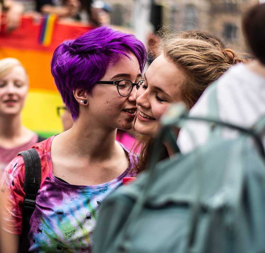 A young person with cropped purple hair kisses their girlfriend on the cheek. In the background a rainbow LGBTIQ flag can be seen.