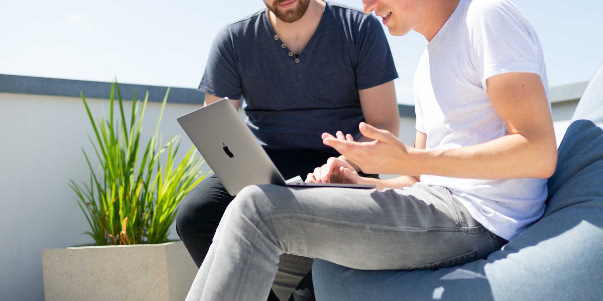 Two men sit outside with one sitting on a beanbag with a laptop on his lap.