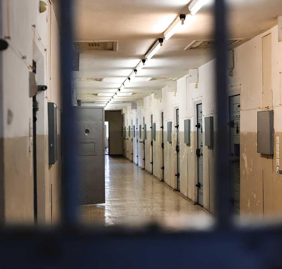 The inside of a prison. A long corridor and one of the cell doors stands ajar.