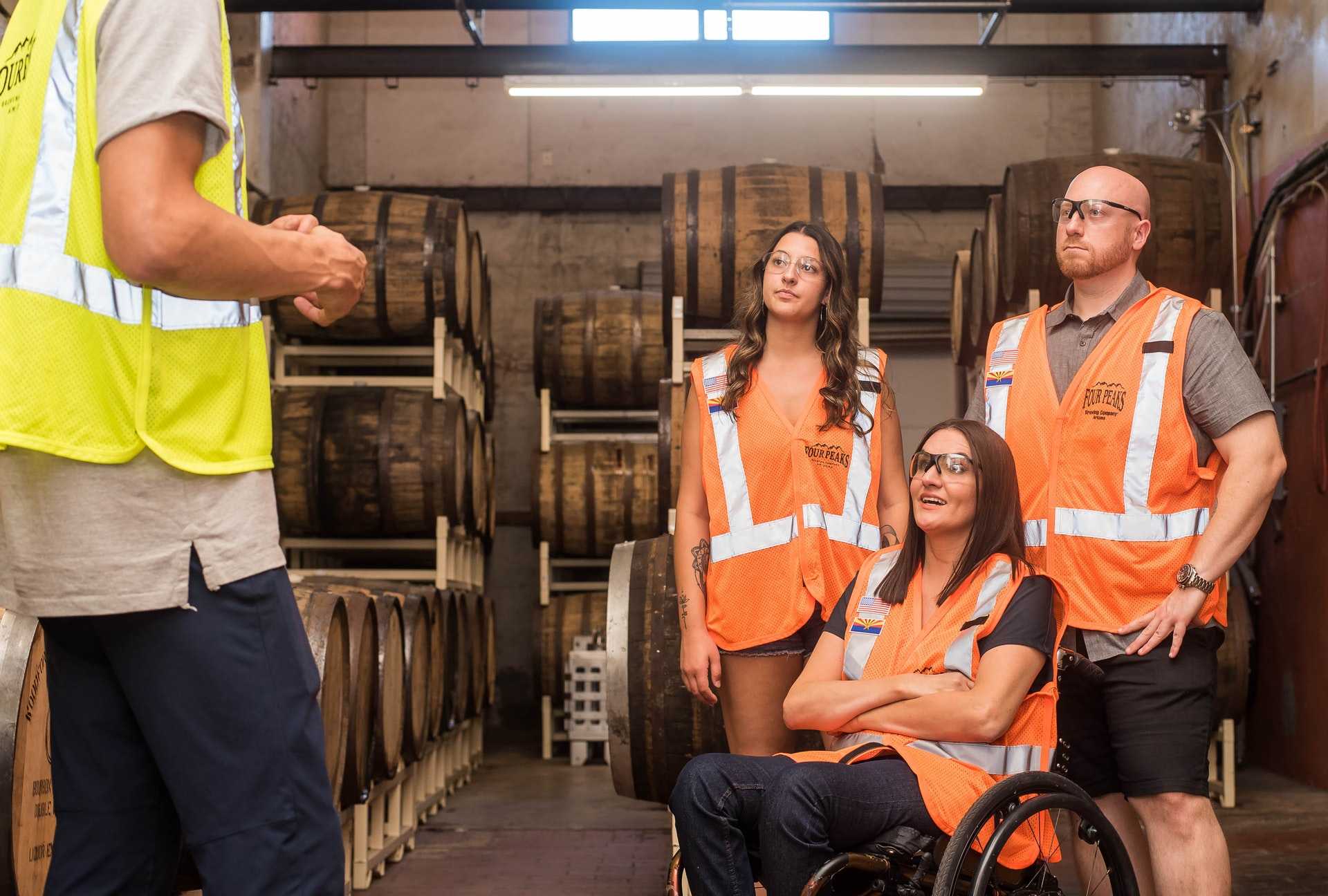 An individual dressed in a yellow vest is speaking a to group dressed in orange vests, including one in a wheelchair in a brewery