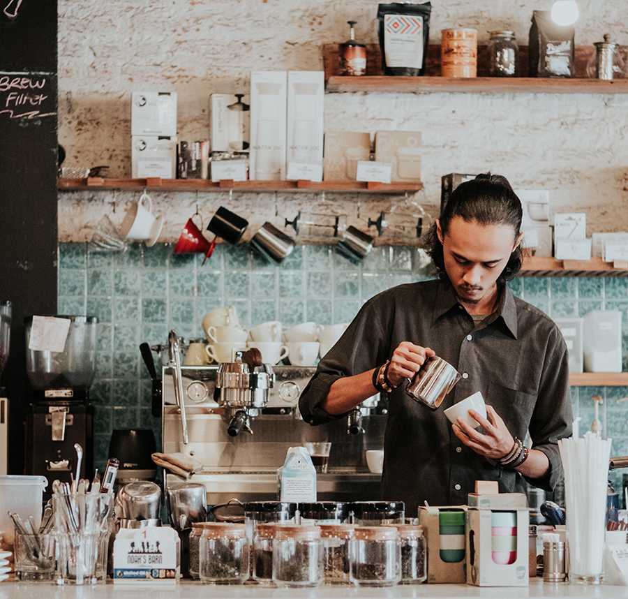 A young barista pours a coffee behind a counter in a cafe.