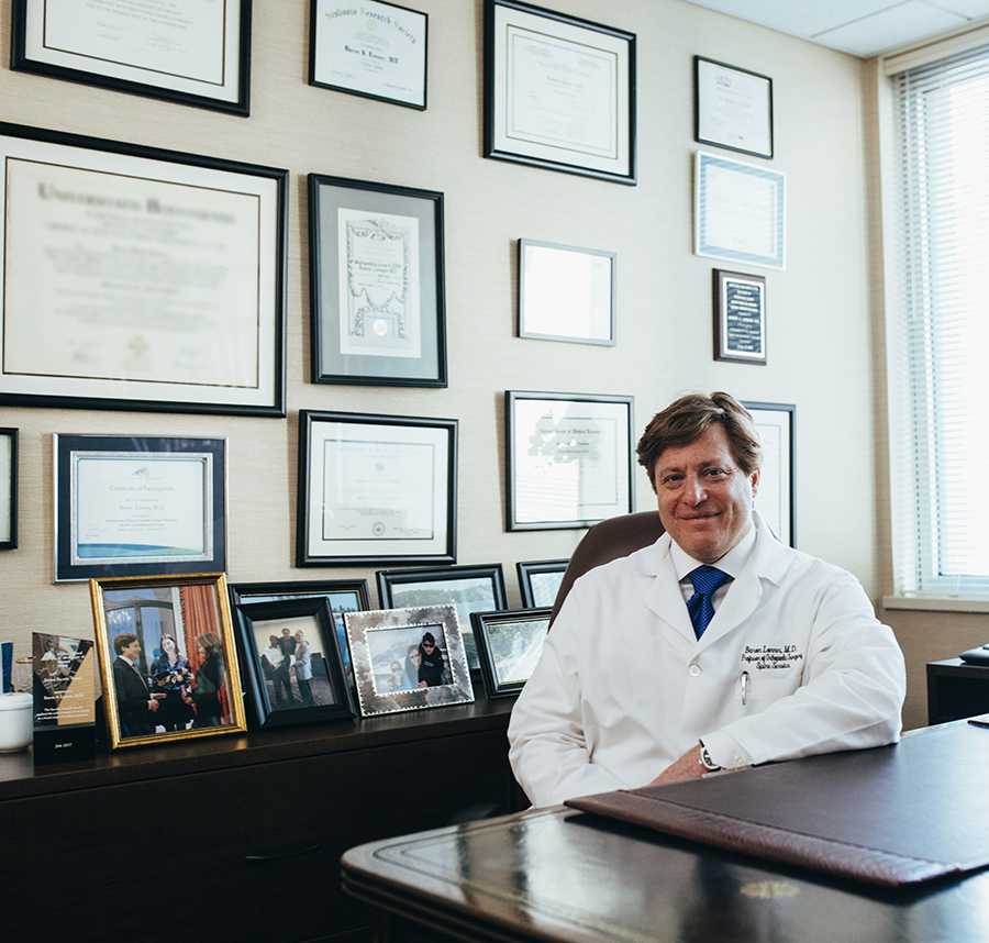 A doctor sits in his office. He is wearing a white coat and his degrees hang framed on the wall behind him.