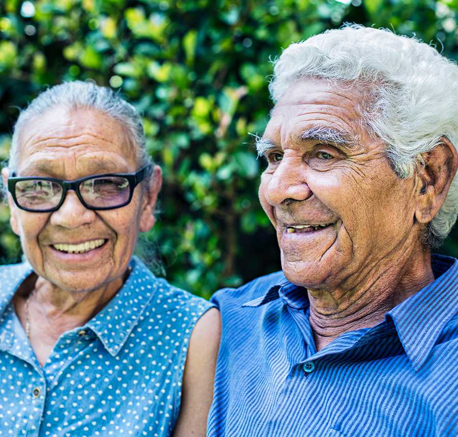 An elderly Aboriginal couple sit smiling in the gardens.