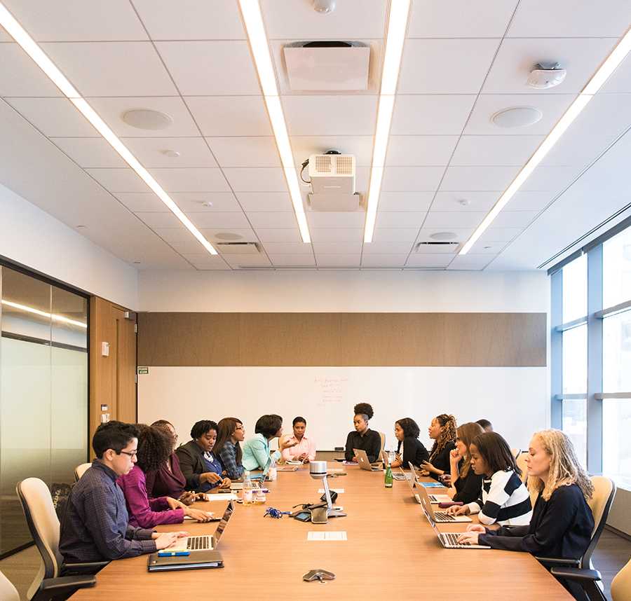 A group of workers in a business meeting. They sit at a long table. They all have their laptops out.