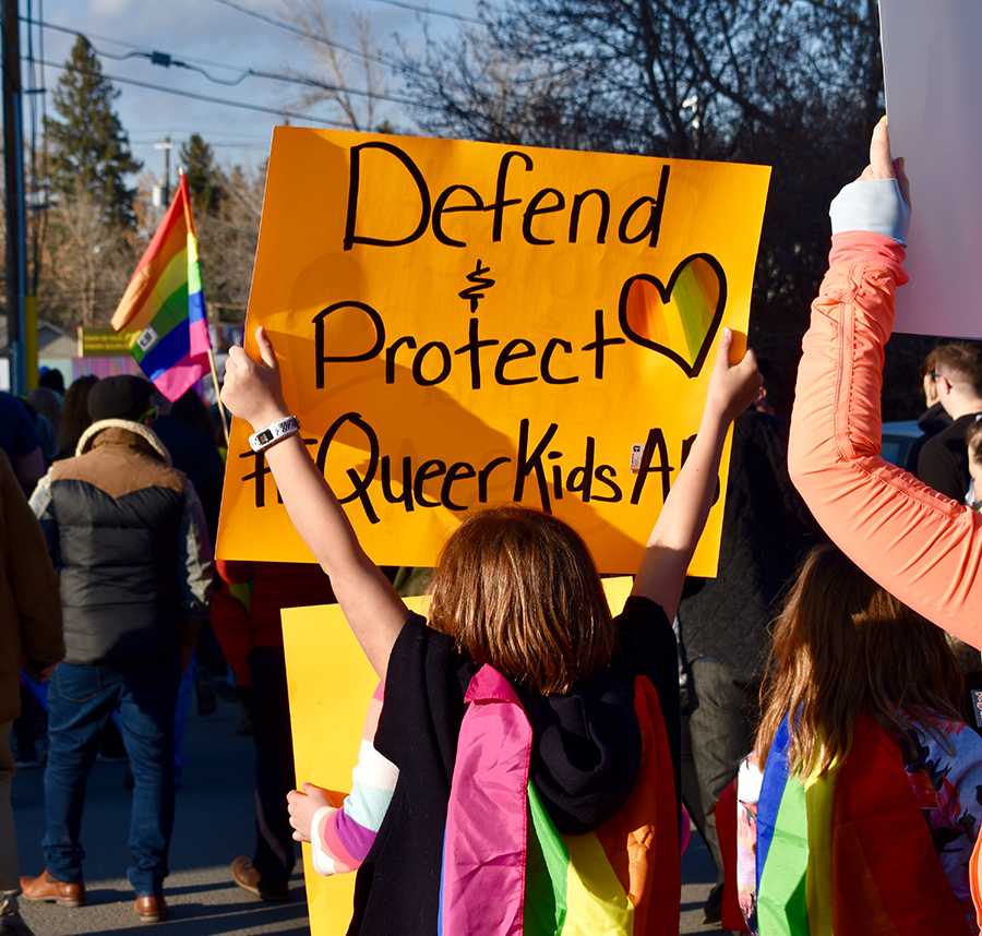 A young child holds up a bright sign at a protest. The sign reads 'defend and protect queer kids'.