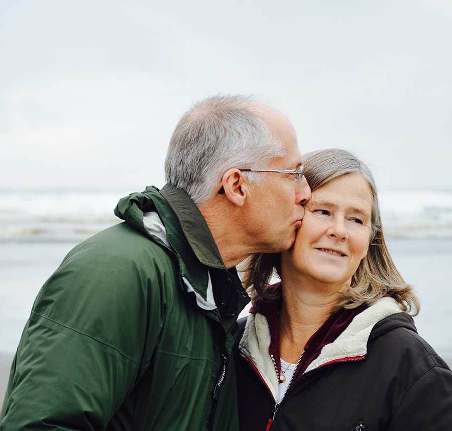 A older couple on the beach. The husband is kissing his wife on the cheek.