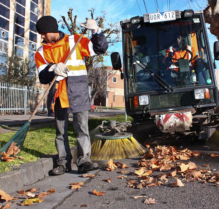 A government employee rakes the leaves in the gutter. Another employee sits in a street sweeper.