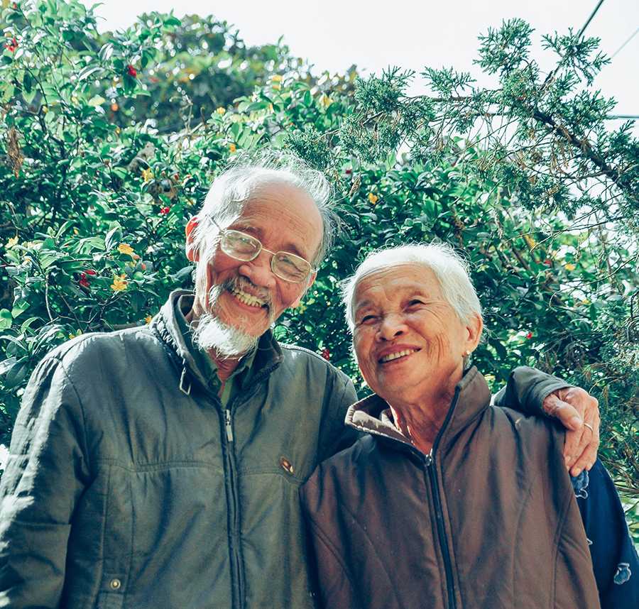 An elderly Asian couple stand smiling with their hands around each other