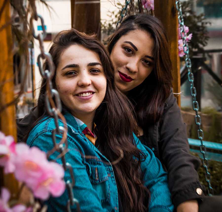 A lesbian couple sit on a swing chair. One of women has her arm around the other.