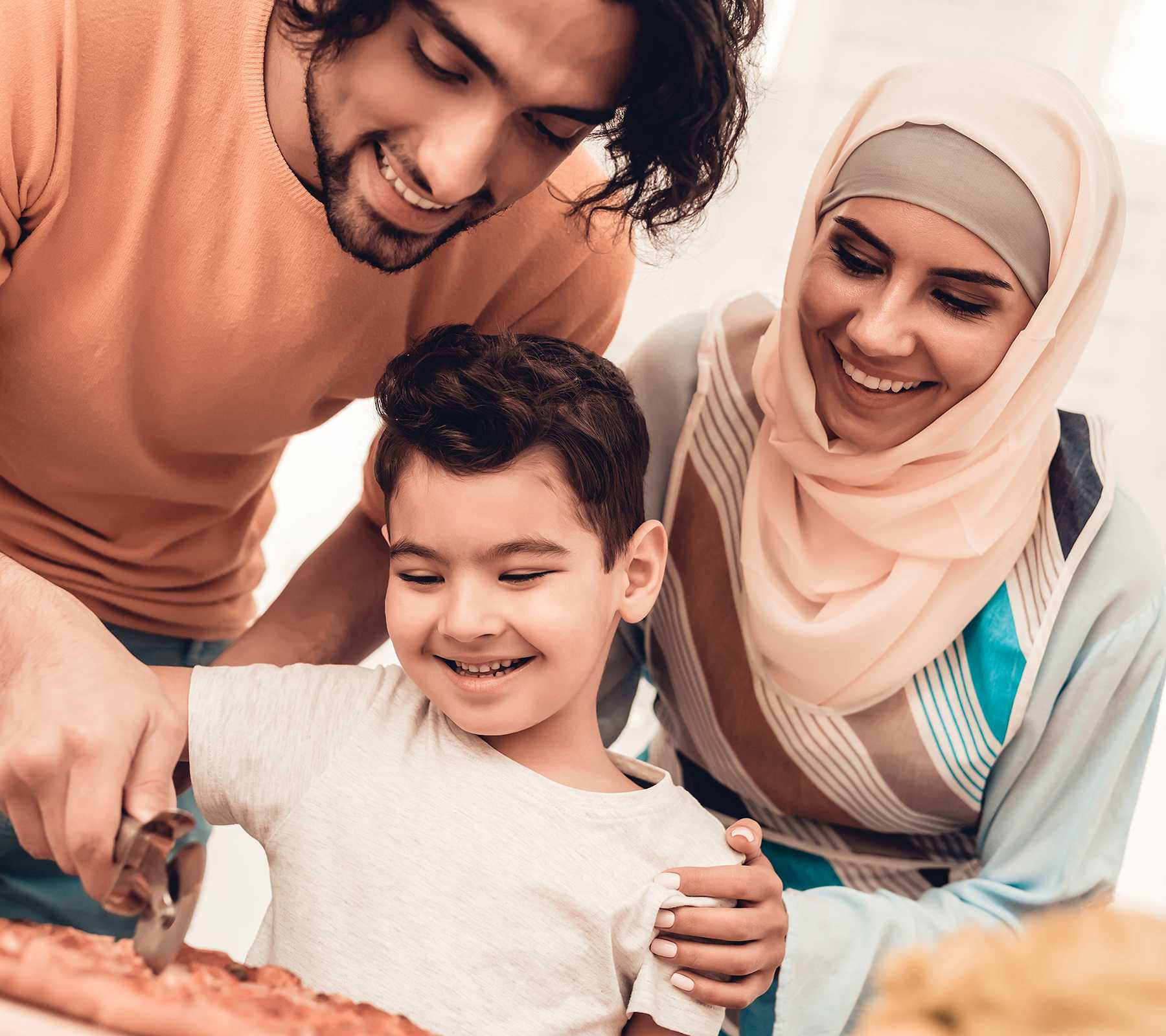 Happy Arabian family eating pizza in kitchen. The father helps the son chop his pizza.