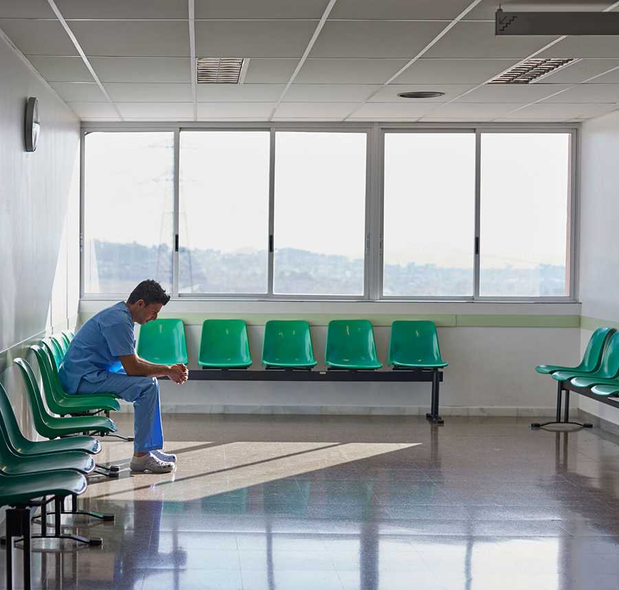 Man in scrubs sits on a plastic chair in an empty waiting room, looking down.