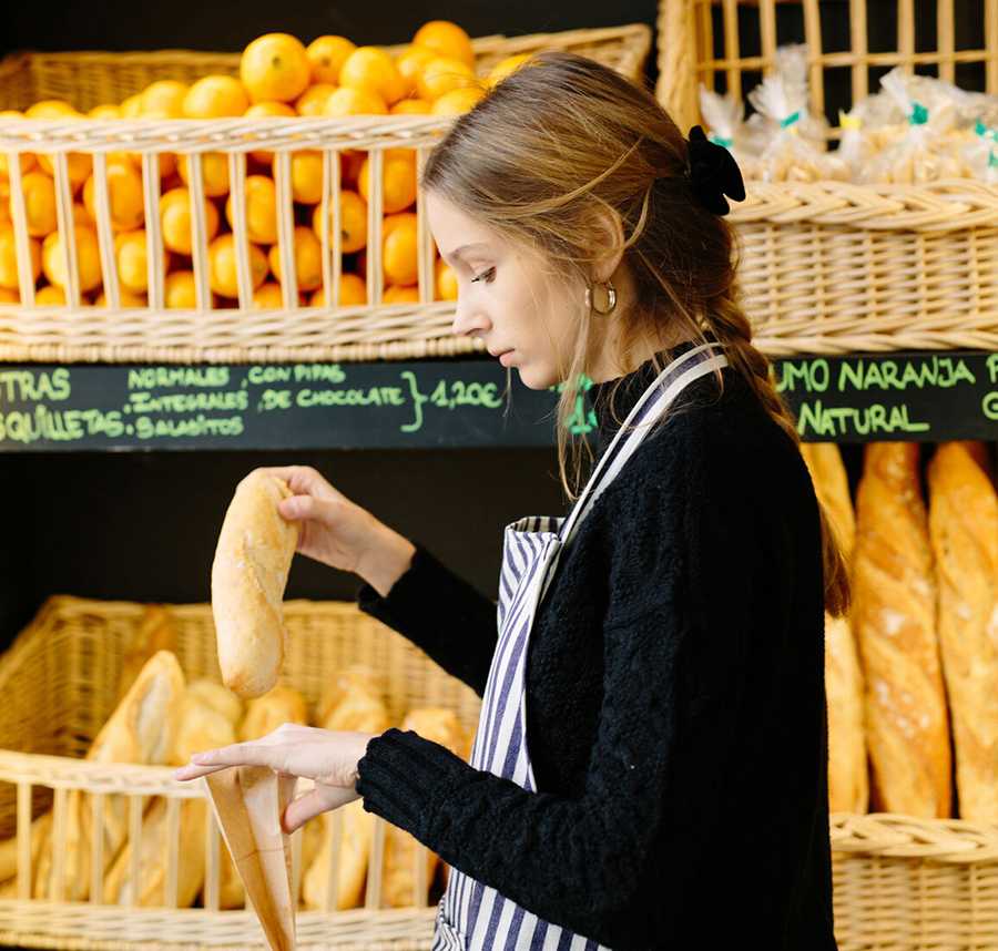 A young bakery worker in an apron places a bread roll into a bag.