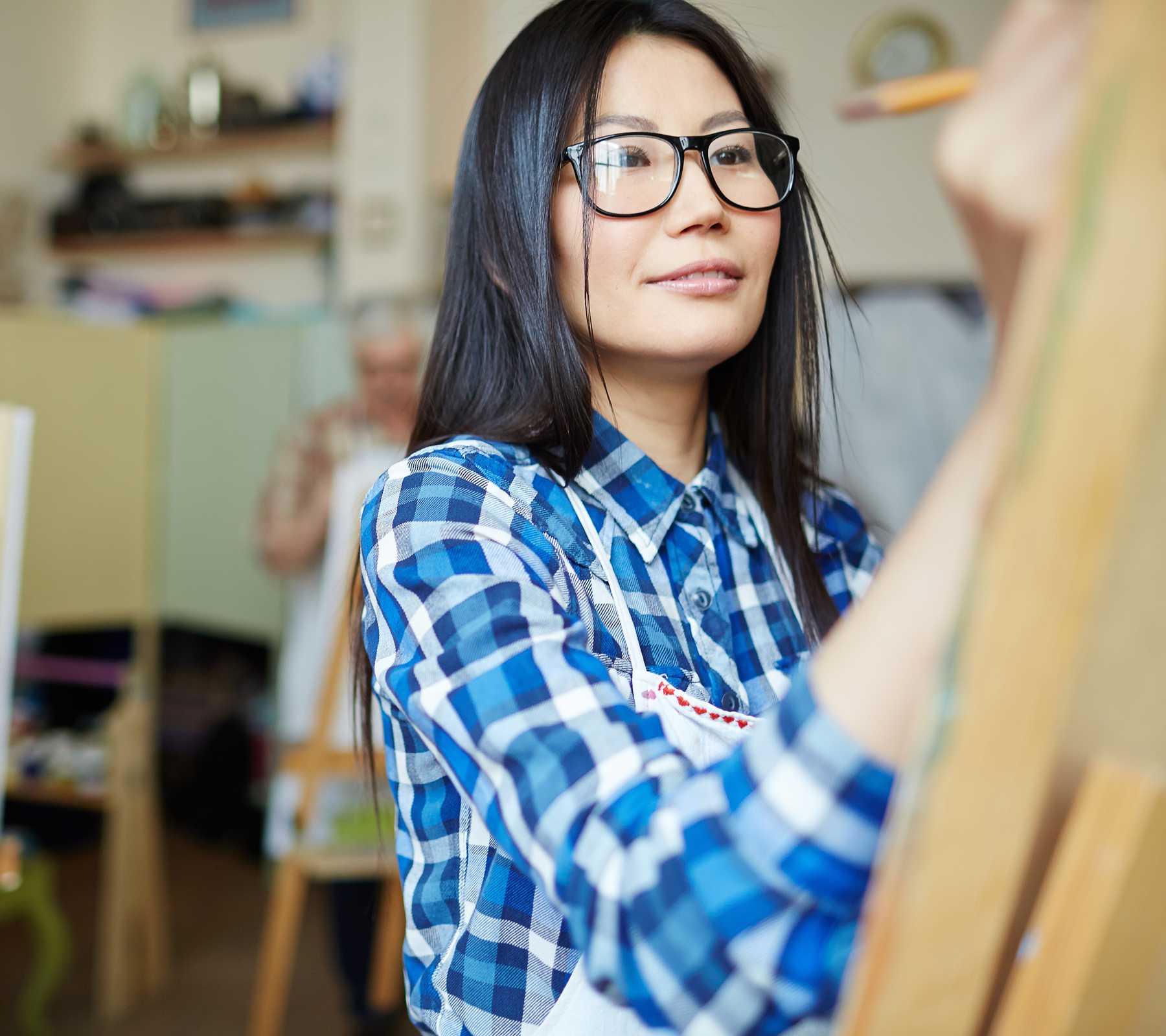 A young woman in a blue checkered shirt holds her hand up to an easel with a brush