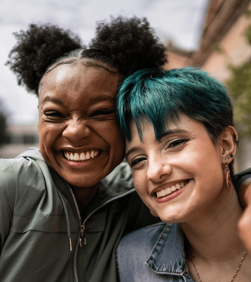 Two young women with their arms around each other smile at the camera. One has curly dark hair, the other a vivid teal green.