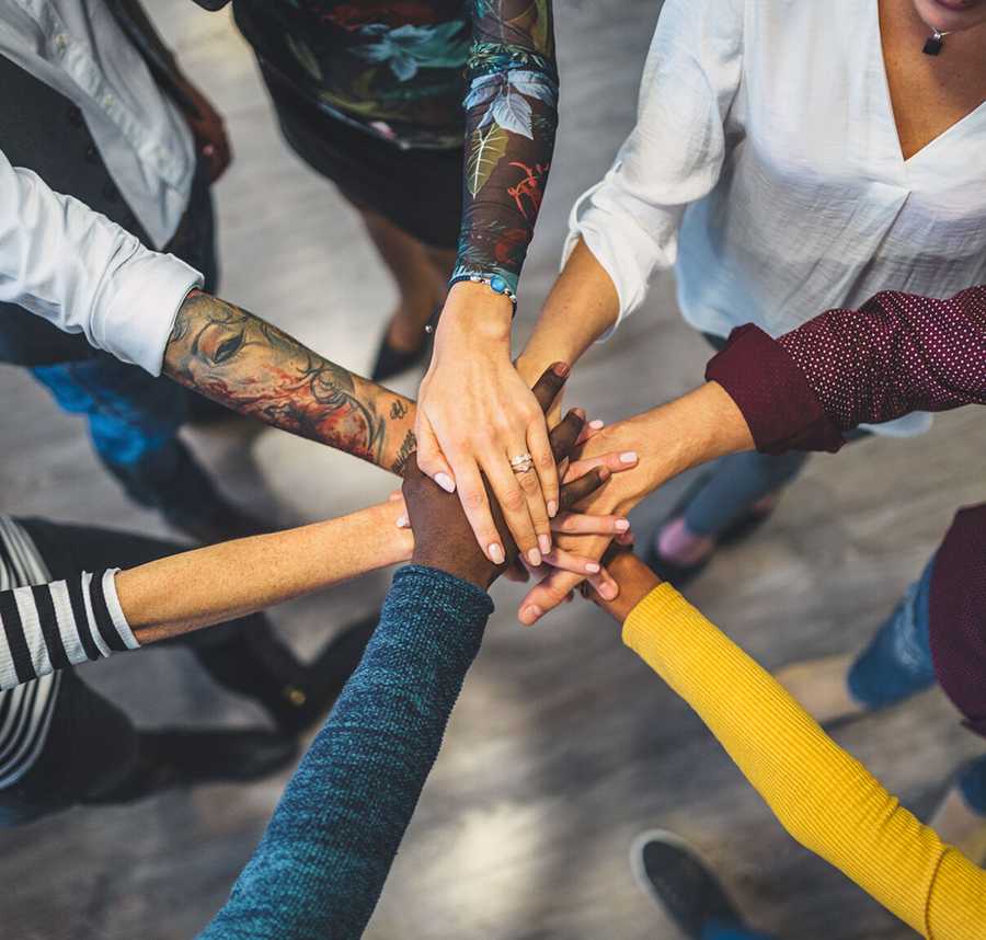 A group of people from different backgrounds and cultures stack their hands on top of each other in a circle.