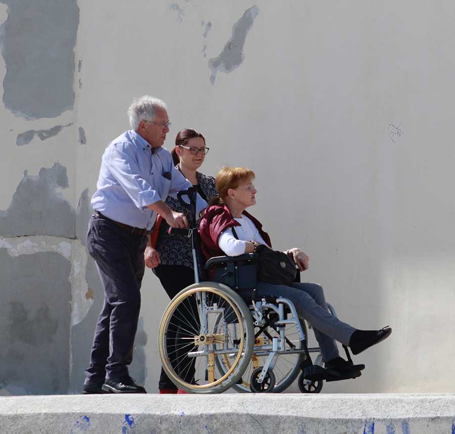 An older man and woman push their friend in a wheelchair.